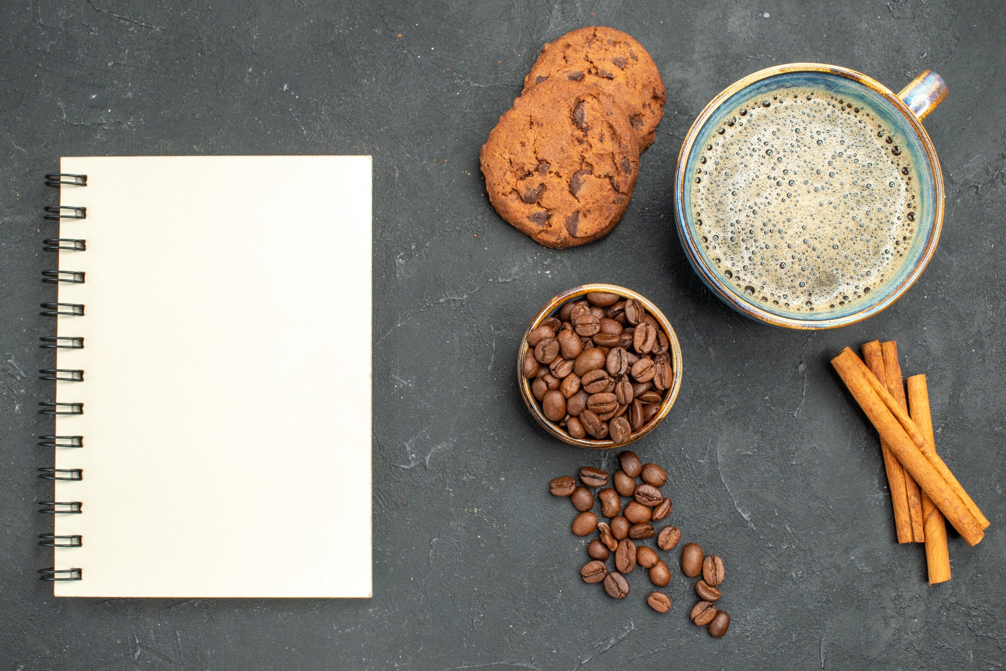 un carnet sur un plan de travail en grès, accompagné d'une tasse de café, d'épices, de cookies et de café en grains.