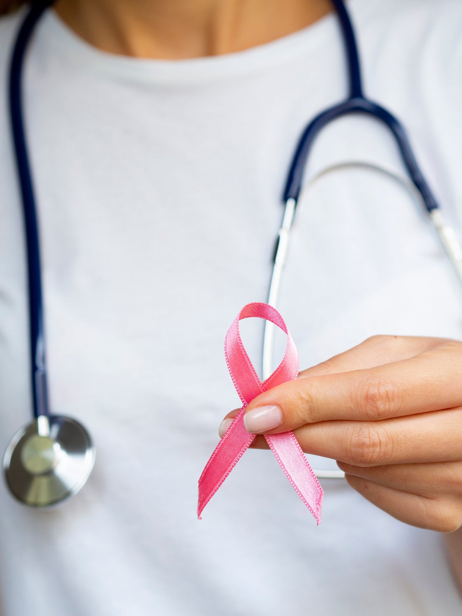 médecin en arrière plan avec un stéthoscope autour du cou, en premier plan sa main qui tient un ruban rose, symbole de la lutte contre les cancers féminins